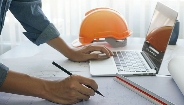 A person planning on an architectural blueprint with a laptop and hard hat nearby.